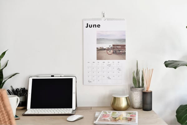 A custom photo wall calendar open to June hangs above a desk with a tablet and plants. A custom photo wall calendar open to June hangs above a desk with a tablet and plants.
