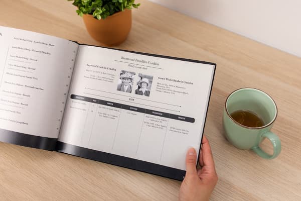 A hand holds open a Family History Book showing a Family Group Sheet for Raymond, his wife and children.