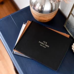 Three Family History Genealogy Books sit on an end table. The top book has a vegan black leather cover and gold title.