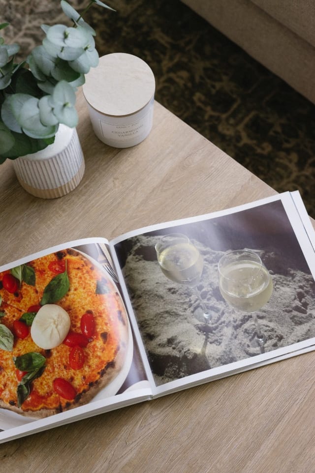 An open Premium Hardcover Photo Book on a wood coffee table, showing a pizza photo and two glasses of white wine. An open Premium Hardcover Photo Book on a wood coffee table, showing a pizza photo and two glasses of white wine.