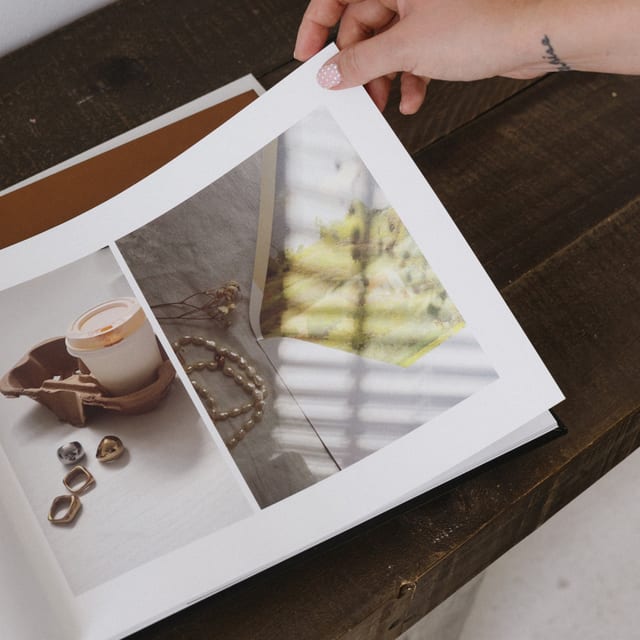A hand turns a page of a Premium Hardcover Photo Book showing still life photos of a candle, pearls and jewelry, on a dark wood table. A hand turns a page of a Premium Hardcover Photo Book showing still life photos of a candle, pearls and jewelry, on a dark wood table.