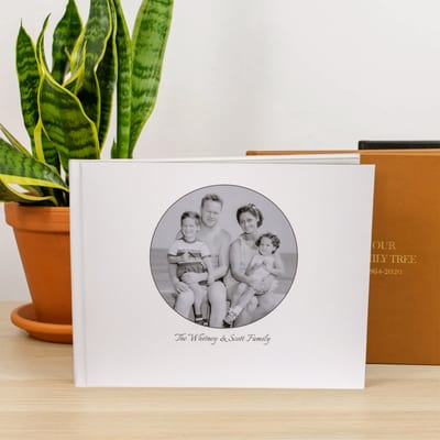 A Family History Book with a custom photo cover, showing a black and white family photo, stands on a wood table beside a houseplant.