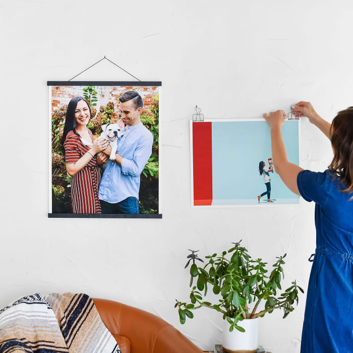 Two Fine Art Prints are hung above a big comfy chair. The first hangs from Black Poster Rails, and the second is being hung from Skeleton Clips by a woman in a blue dress.