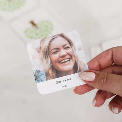 A hand holds a MyCanvas Family Flip memory game card showing a photo of Grace Kent, above blurred tree-backed cards.