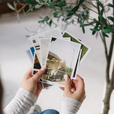 Two hands hold a fanned set of Glossy Photo Prints of travel and outdoor scenes, beside an olive plant.