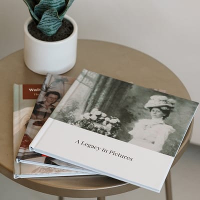 Two MyCanvas Hardcover Photo Books rest on a round side table beside a potted snake plant, the top one titled A Legacy in Pictures with a vintage portrait of a woman in a wide hat.