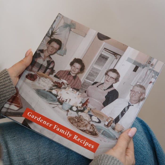 Hands hold a MyCanvas Hardcover Photo Book titled Gardener Family Recipes, its cover showing a vintage photo of a family gathered around a dinner table.