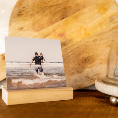 A square metal Desk Print of a father carrying his child into ocean waves, propped on a wood base stand. A square metal Desk Print of a father carrying his child into ocean waves, propped on a wood base stand.