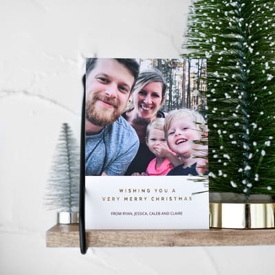 A Holiday Card is propped up on a wooden shelf next to several miniature Christmas trees. A Holiday Card is propped up on a wooden shelf next to several miniature Christmas trees.
