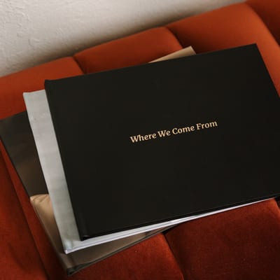 A stack of MyCanvas Premium Hardcover Photo Books rests on a rust velvet couch, the top one in black linen with a gold foil title reading Where We Come From.