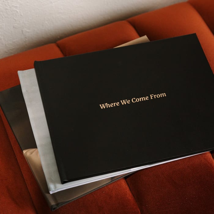 A stack of MyCanvas Premium Hardcover Photo Books rests on a rust velvet couch, the top one in black linen with a gold foil title reading Where We Come From.