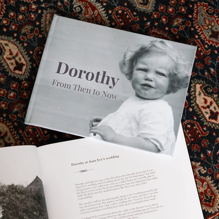A MyCanvas Premium Hardcover Photo Book titled Dorothy From Then to Now lies open on a patterned rug, its cover showing a vintage black and white portrait of a young child.