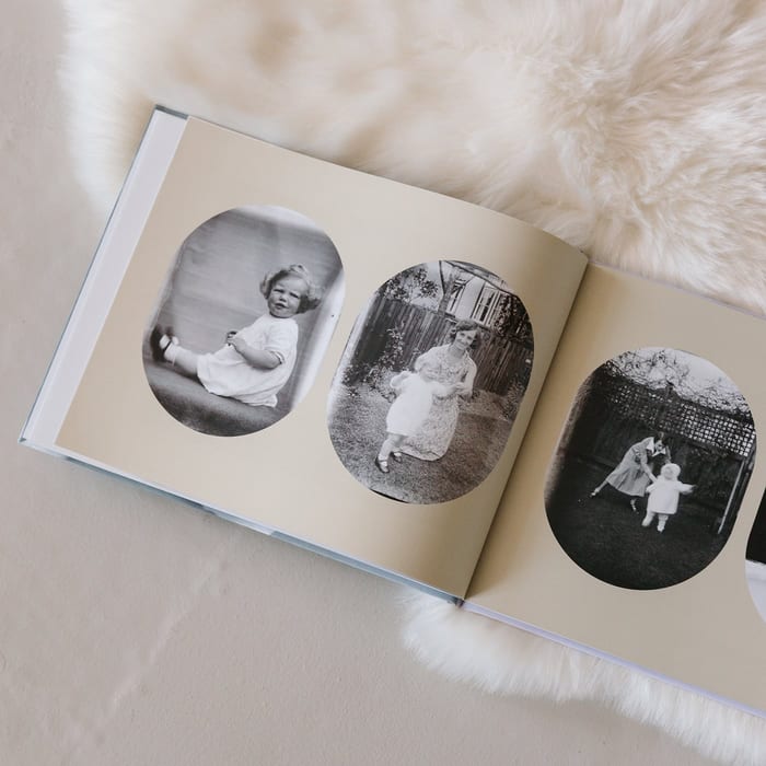 A MyCanvas Premium Hardcover Photo Book lies open on a sheepskin rug, showing a spread of vintage black and white family photos in oval frames.