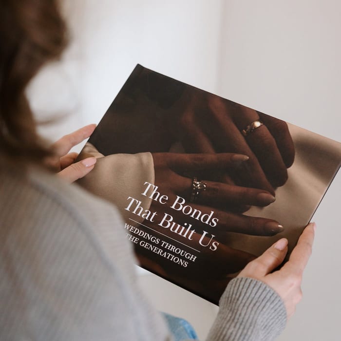 A woman holds a MyCanvas Premium Hardcover Photo Book titled The Bonds That Built Us, Weddings Through the Generations, its cover showing close-up of clasped hands with rings.