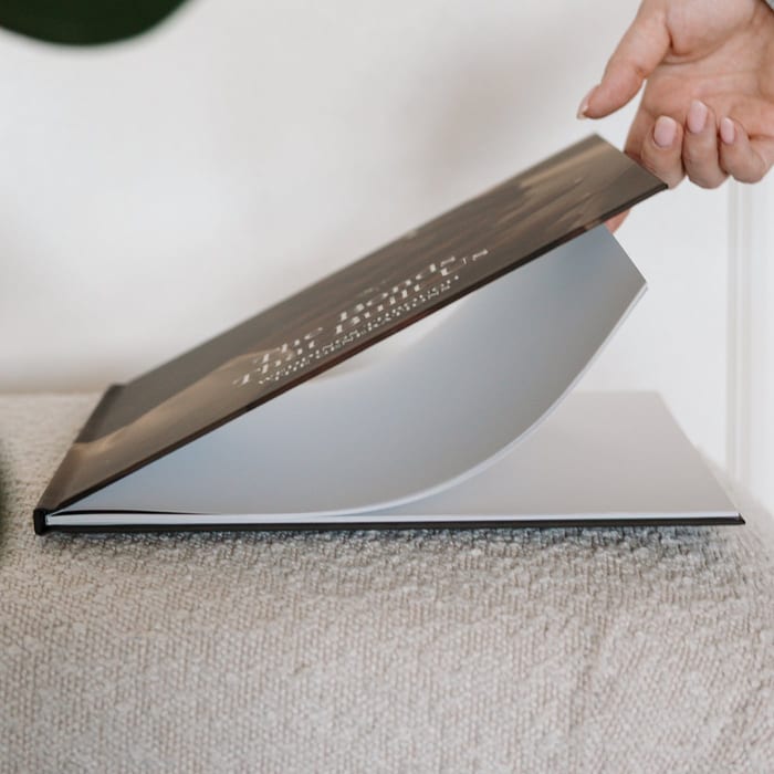 A hand lifts the lay-flat cover of a MyCanvas Premium Hardcover Photo Book, showing its sturdy spine and thick pages.