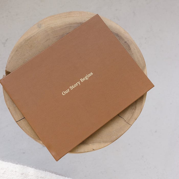 A MyCanvas Premium Hardcover Photo Book in tan linen with a gold foil title reading Our Story Begins sits on a round wooden tray.