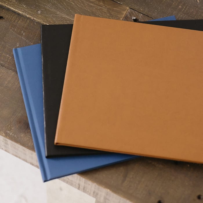 Three MyCanvas Premium Hardcover Photo Books in tan, black, and blue linen covers are fanned out on a rustic wood surface.