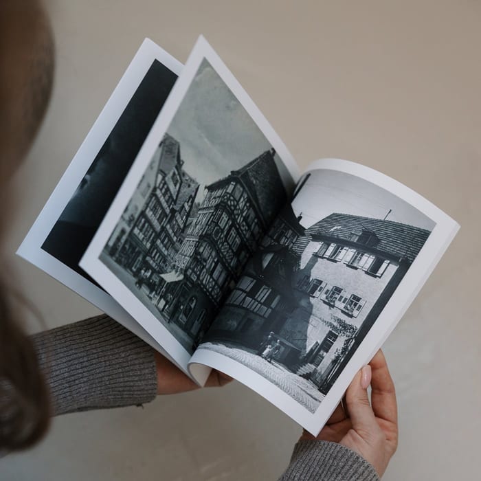 A person flips through a MyCanvas Softcover Photo Book, showing a black and white spread of old half-timbered houses on a town street.