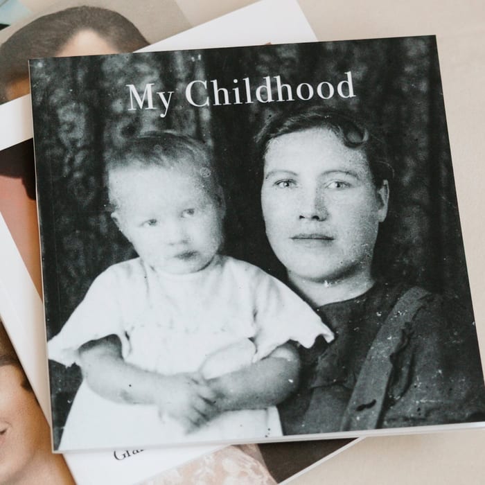 A MyCanvas Softcover Photo Book titled My Childhood rests on a table, its cover showing a vintage black and white portrait of a mother holding a baby.