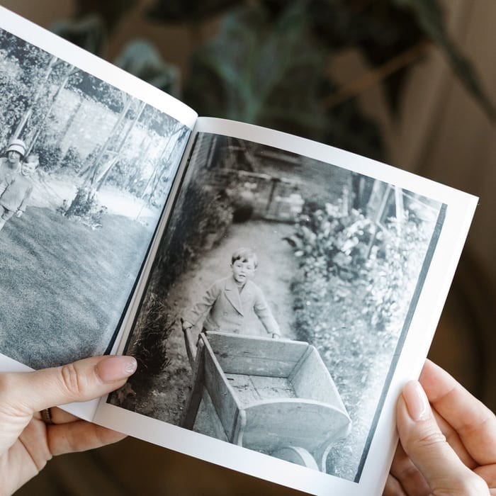 Two hands hold open a MyCanvas Softcover Photo Book to a black and white spread of a young child pushing a wooden cart in a garden.