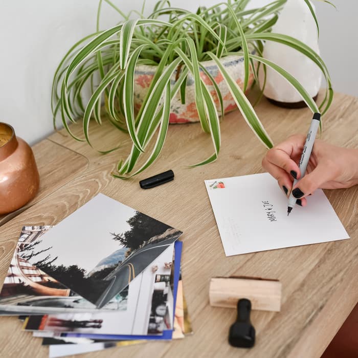 A person addresses a Square white Envelope next to a stack of vacation photos.
