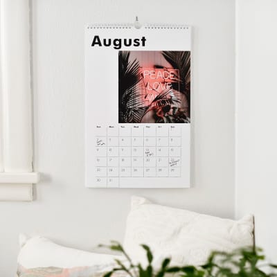 A Large custom photo Wall Calendar showing a photo, August, with events and birthdays filled in, hangs above a windowseat.